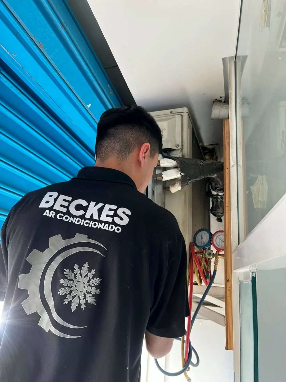 Técnico de costas com a polo preta da Beckes Ar Condicionado, trabalhando em uma evaporadora com o manifold conectado