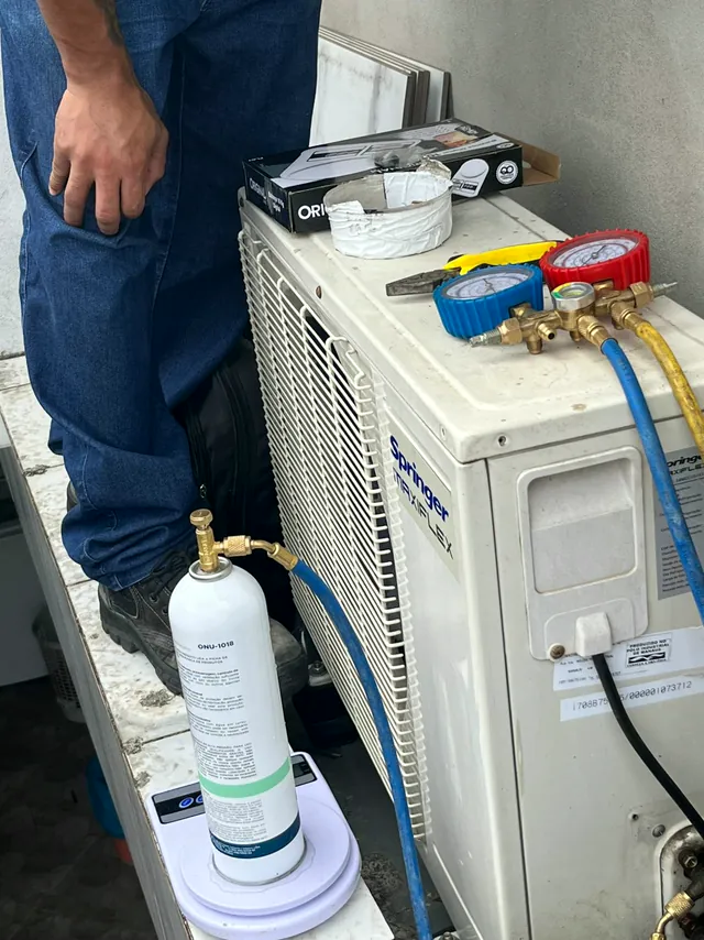 Cilindro de gás refrigerante sobre uma balança, ligado por mangueira à condensadora, com manifold sobre o aparelho